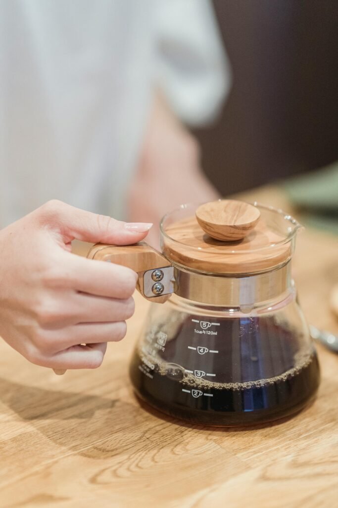 A hand holds a glass pot filled with freshly brewed black coffee on a wooden table.